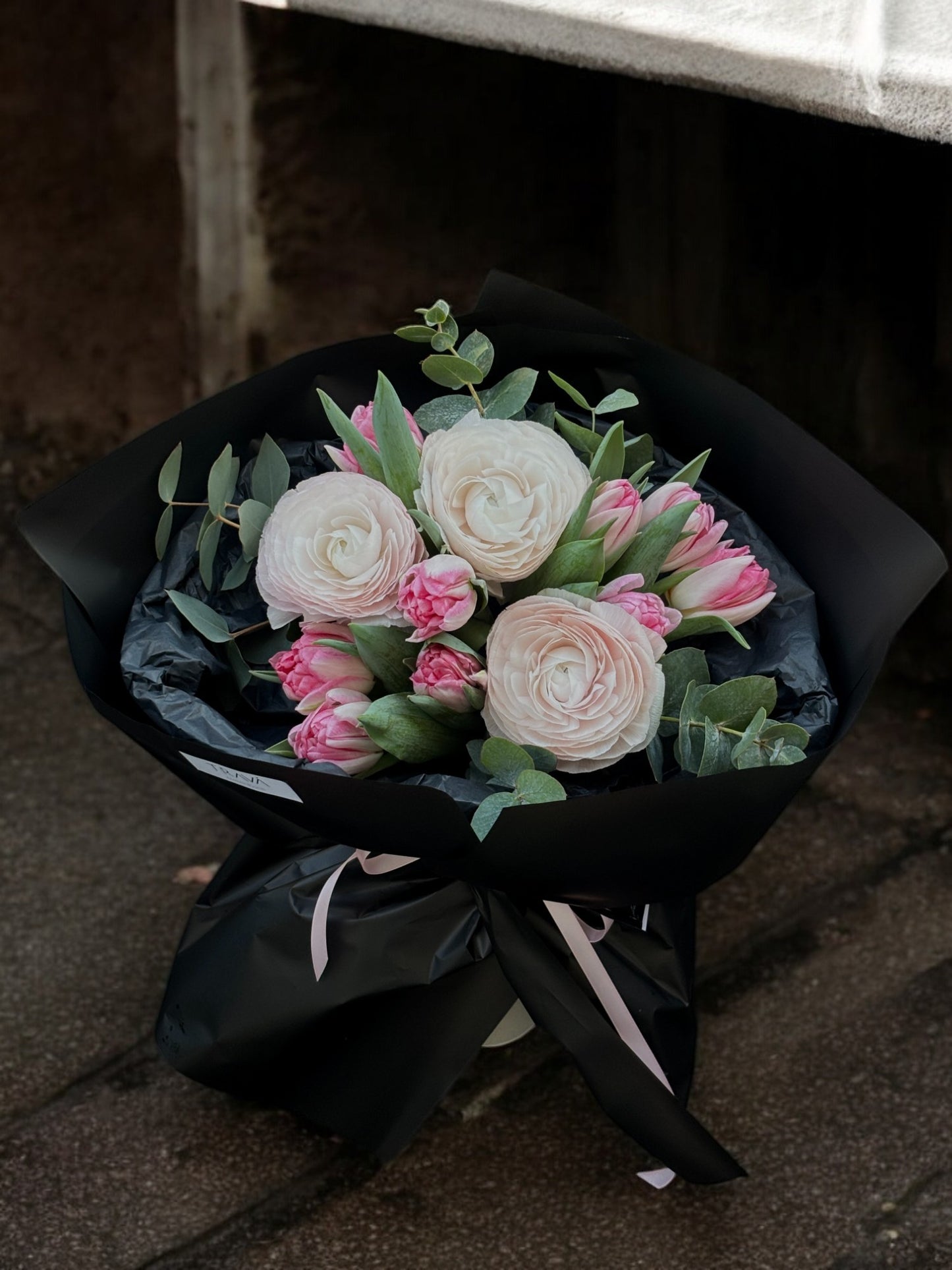 Buchet elegant cu ranunculus alb, lalele roz și eucalipt, ambalat în hârtie neagră, perfect pentru livrare la domiciliu în Oradea.
