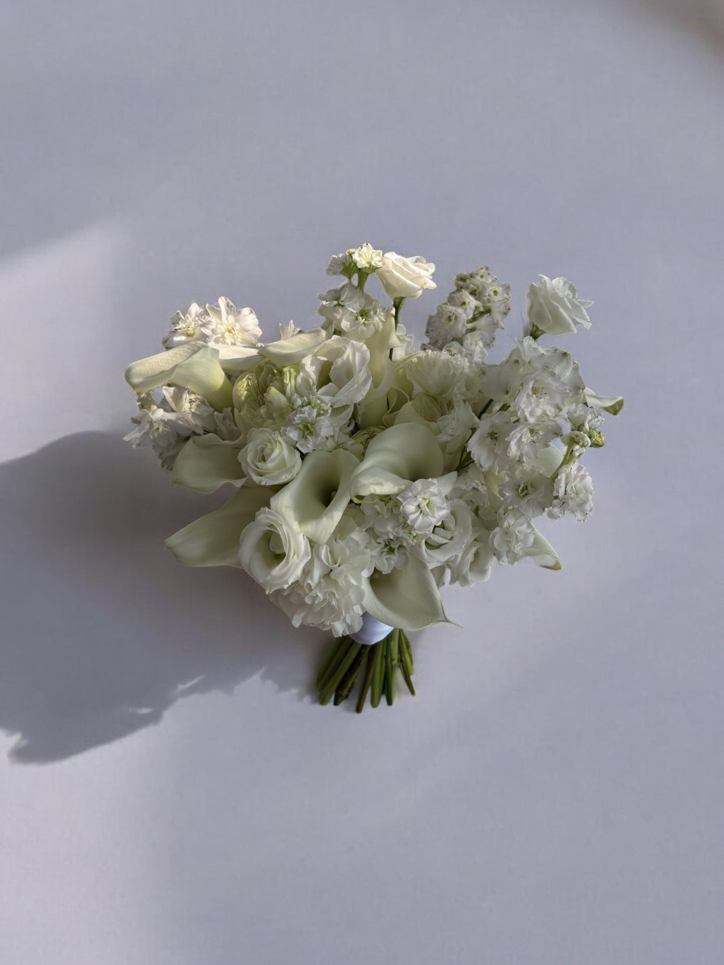 Buchet de mireasă din flori albe, cu cale, delphinium, trandafiri, lisianthus și garoafe, perfect pentru nunți și ocazii speciale.