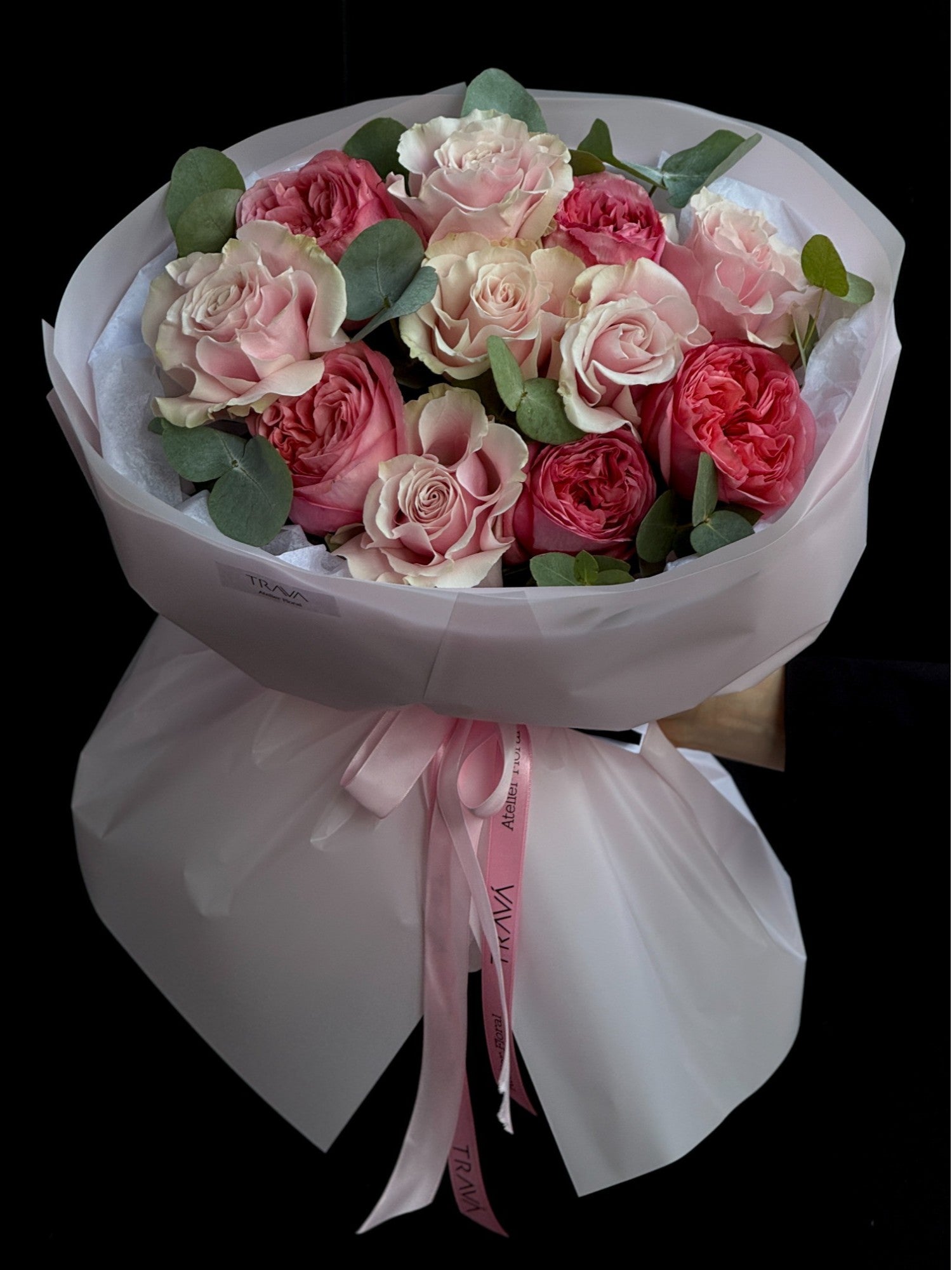 Un buchet de trandafiri roz și coral, cu frunze de eucalipt, ambalat în hârtie albă și panglică roz, oferit de florărie Oradea TRAVÁ cu livrare flori la domiciliu.