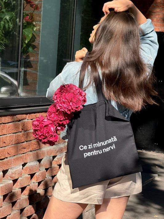 Sacoșă din bumbac negru cu mesaj Ce zi minunată pentru nervi, purtată ca tote bag cu flori.