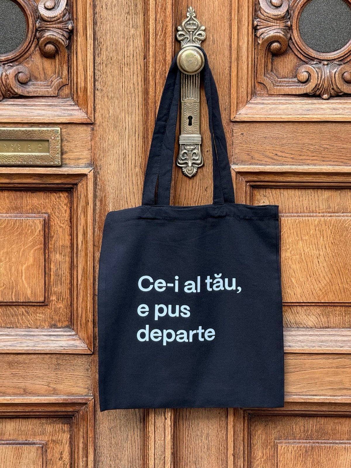 Tote bag din bumbac negru 100%, cu mesaj Ce-i al tău, e pus departe, atârnată de clanța unei uși vintage.