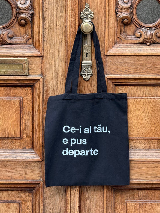 Tote bag din bumbac negru 100%, cu mesaj Ce-i al tău, e pus departe, atârnată de clanța unei uși vintage.