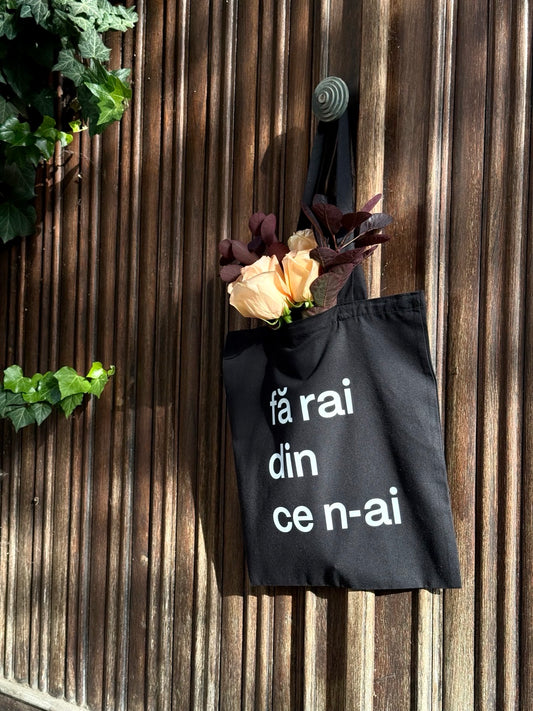 Tote bag din bumbac negru cu mesaj fă rai din ce n-ai, surprins pe fundal rustic din lemn, accesoriu eco și practic.