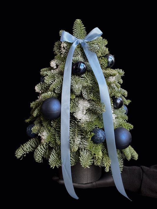 Bradut de Craciun din brad natural decorat cu globuri albastre si funda de satin, tinut in mana, realizat de Trava Atelier Oradea.