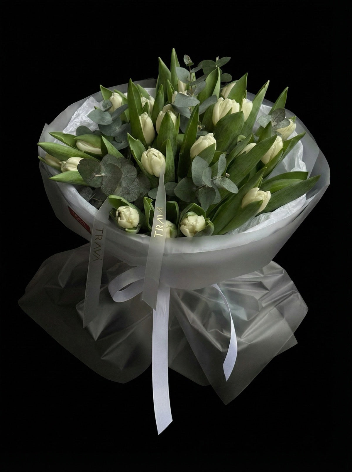 Buchet format din lalele albe și frunze de eucalipt ambalat în hârtie albă mată și legat cu panglică personalizată TRAVÁ.