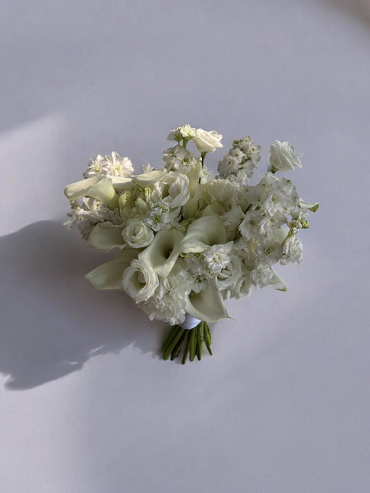 Buchet de mireasă din flori albe, cu cale, delphinium, trandafiri, lisianthus și garoafe, perfect pentru nunți și ocazii speciale.